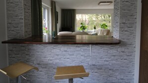 A modern kitchen with a stone-textured backsplash, a wooden countertop, and a view of a living room with a sofa and plants.