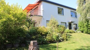 A modern two-story house with a balcony, surrounded by lush greenery and a well-maintained lawn.