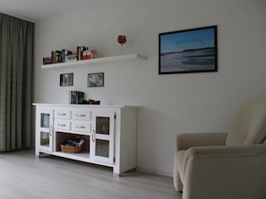 Un salon avec un meuble blanc, un fauteuil beige, une bibliothèque remplie de livres et un tableau encadré accroché au mur.