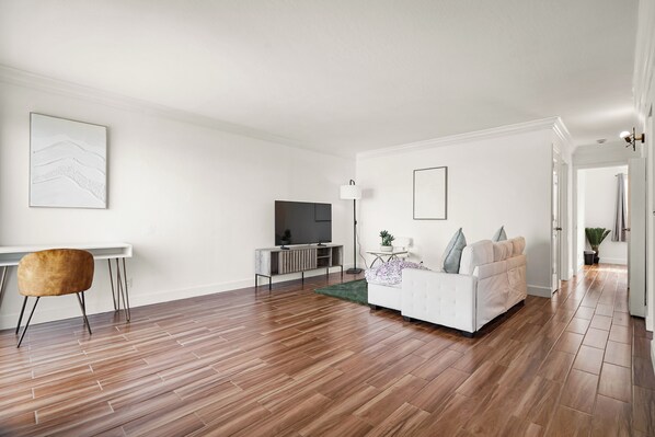 A modern living room with a white sofa, a TV, and a dining area with a chair and table. - Stunning 2BR Ocean Mountain Sunset View (Pacifica)