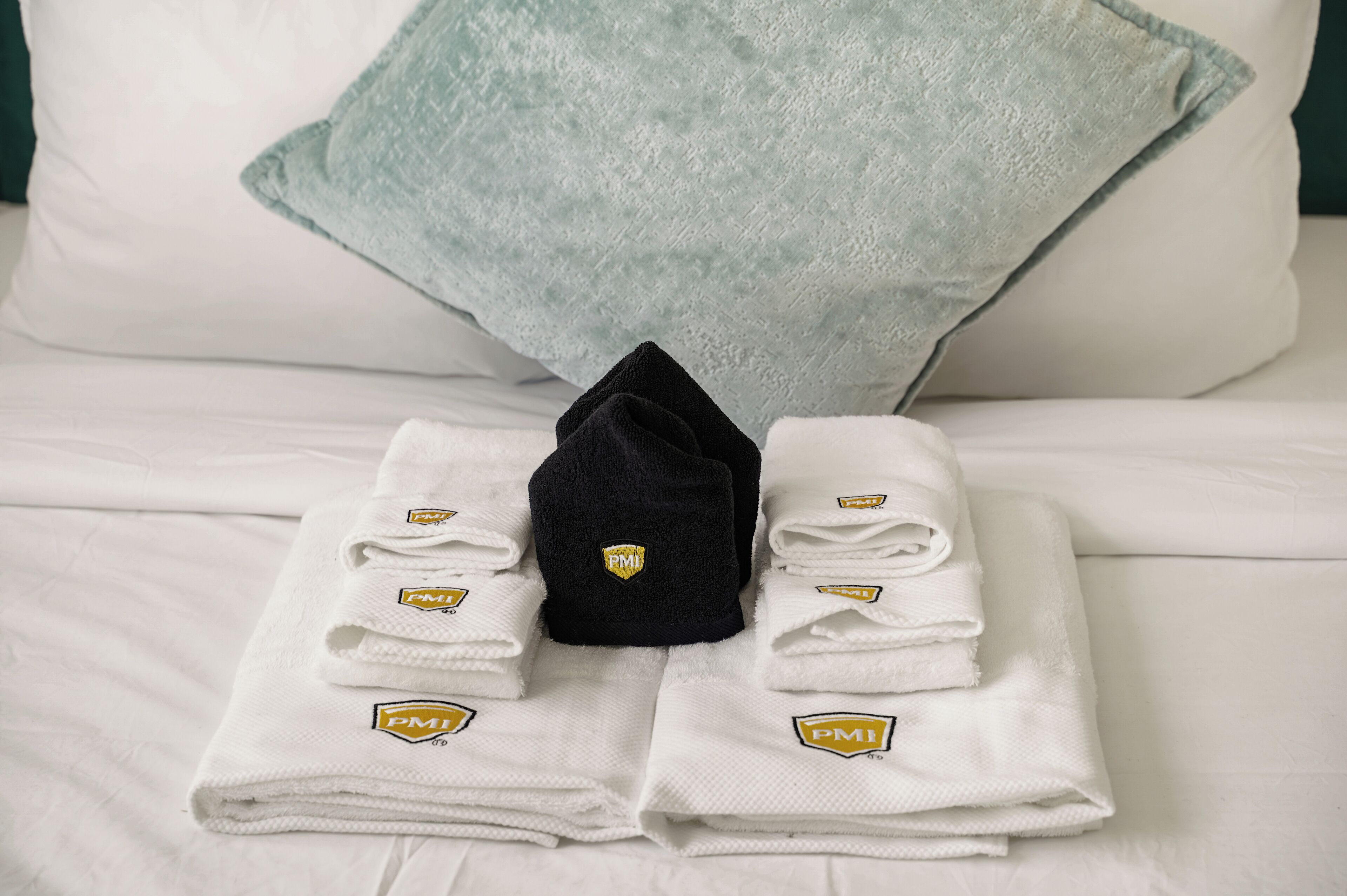 A neatly made bed with white linens, a folded black towel, and a light blue pillow.