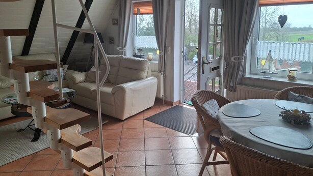 A cozy room with a staircase, a sofa, a dining table with chairs, and a view of the outdoors.