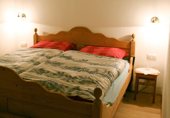 A wooden bed with a floral bedspread, two red pillows, and a bedside table with a lamp.