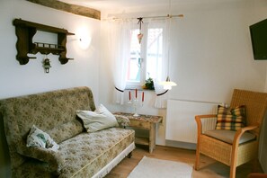 Een kamer met een bank met bloemenprint, een rieten stoel, een tafeltje met een lamp en een raam met gordijnen.