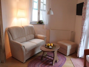 Un salon avec un canapé beige, une petite table ronde sur laquelle se trouve un plateau de fruits, une lampe et un radiateur.