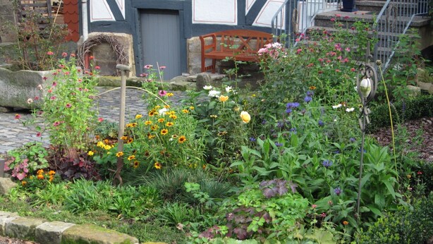 A garden with a variety of flowers and plants, a stone bench, and a building with a dark facade and white trim. - Apartment 'Kammer' with Shared Terrace, Garden, and Wi-Fi (Gudensberg)