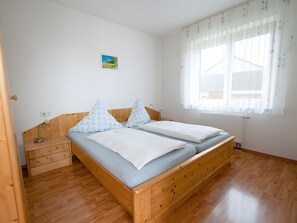 A double bed with a wooden frame, a bedside table, a nightstand, a window with sheer curtains, and a small wall-mounted picture.