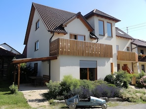 A two-story house with a balcony, a small car, and a garden with various plants.