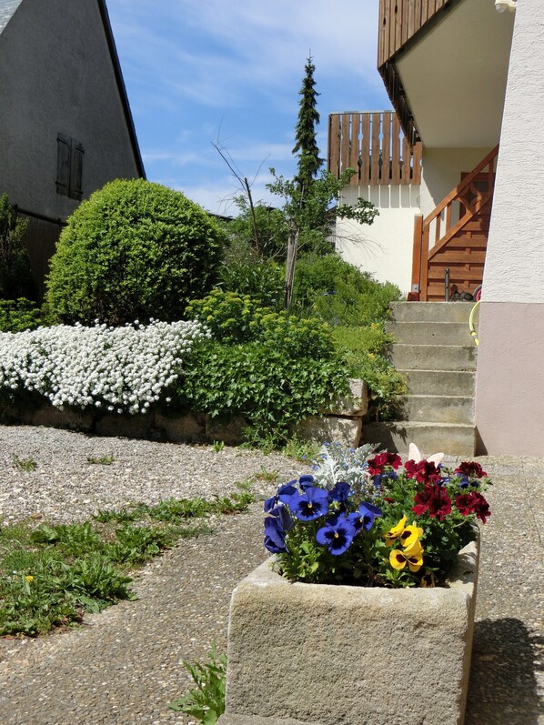 A stone planter with flowers, a gravel path, a wooden staircase, and a building with a sloped roof.