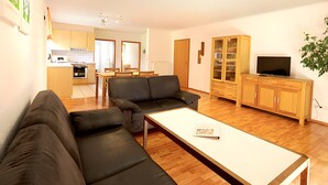 Een moderne woonkamer met een bank, een salontafel en een televisie. Achtergrond is een keuken met houten kasten en een eethoek.