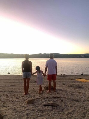 Une famille de trois personnes se tient sur une plage de sable, face à une étendue d’eau calme, avec un coucher de soleil en arrière-plan.
