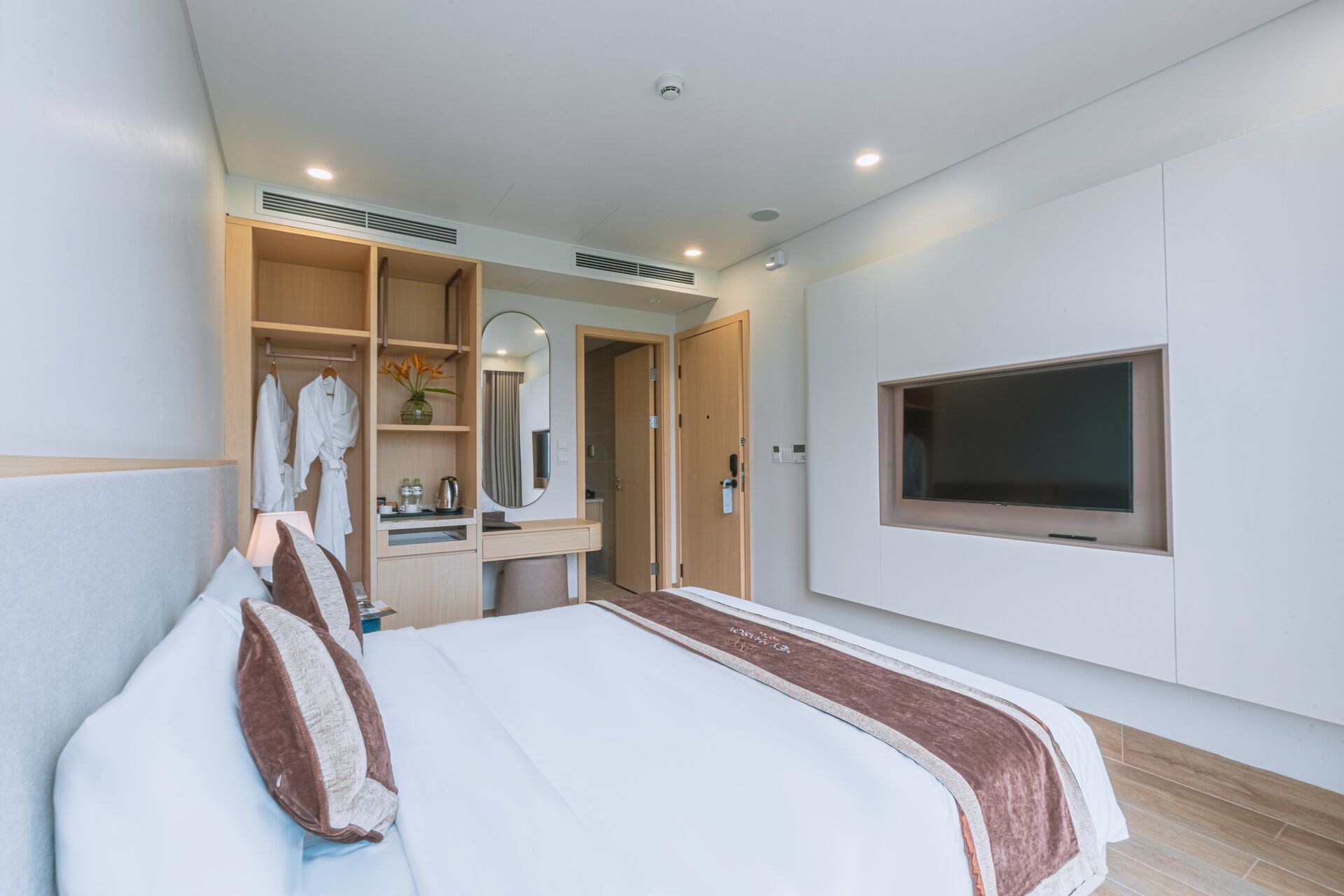 A modern hotel room with a large bed, a vanity with a mirror, a TV mounted on the wall, and a walk-in closet.