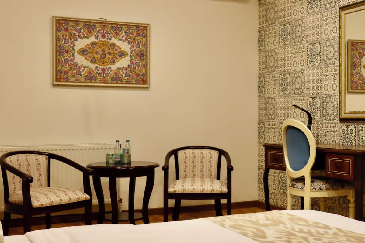 A hotel room with a bed, two chairs, a small table with bottles, an air conditioner, and patterned wallpaper.