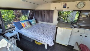 Interno di un camper compatto con un letto, angolo cottura e un piccolo tavolo con una ventola.