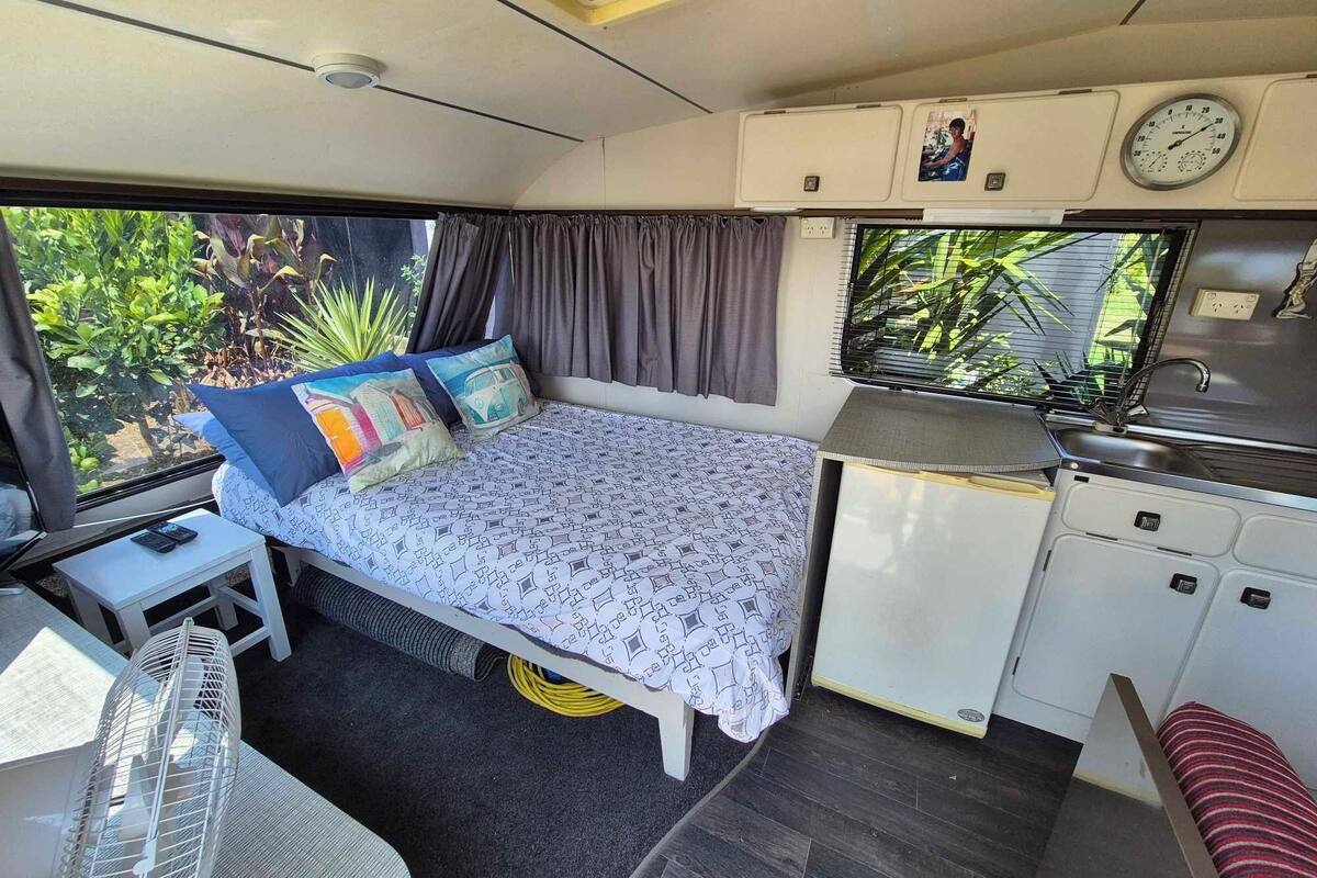 Ein kompaktes Wohnmobil-Interieur mit einem Bett, einer Küchenzeile und einem kleinen Tisch mit einem Ventilator.