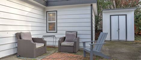 Un espace patio avec deux chaises en osier et une table, un abri de jardin et une maison aux murs recouverts de bardage blanc et dotée d’un toit foncé.