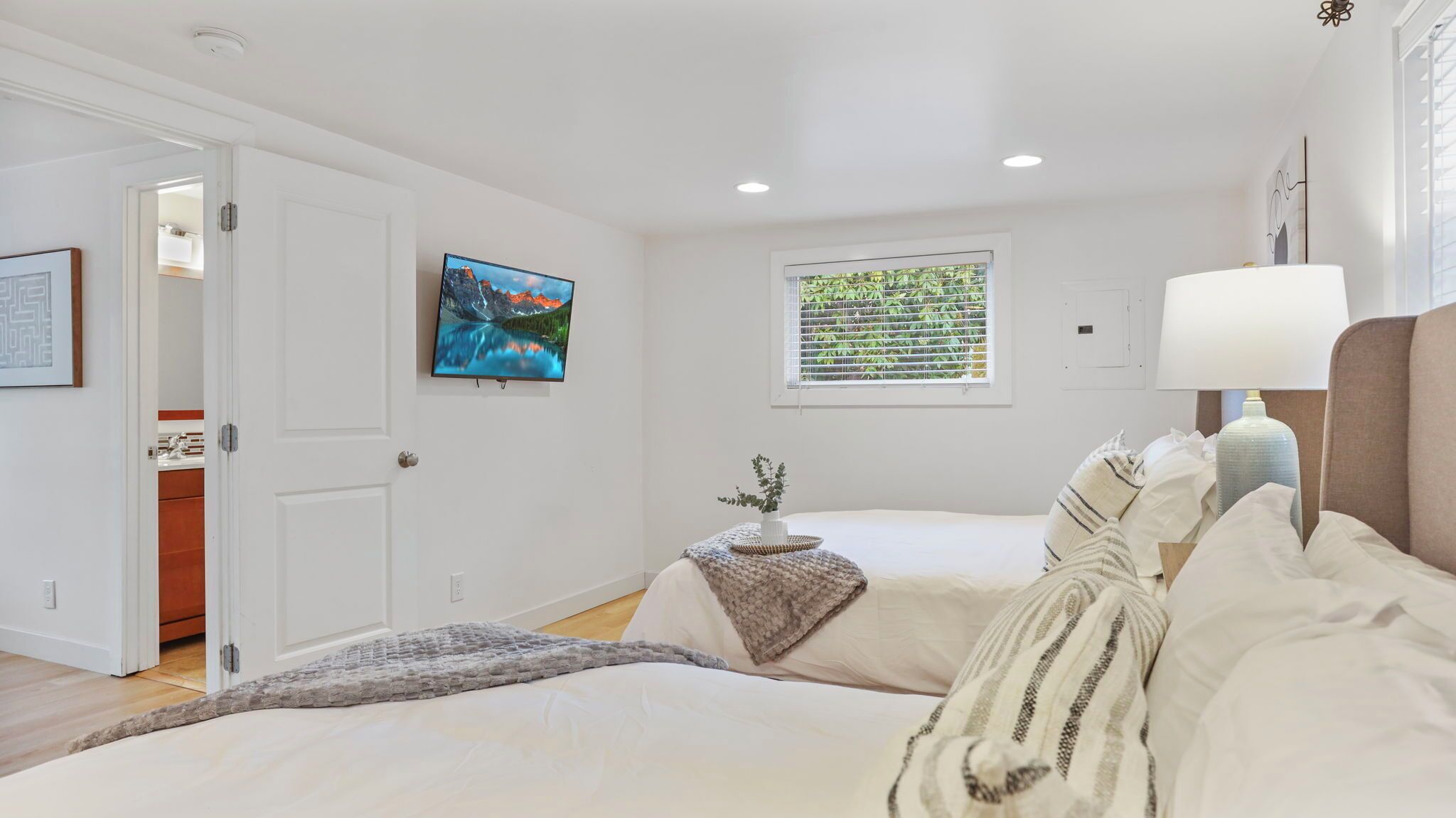 A bedroom with two beds, a TV, a window with blinds, and a bedside lamp.