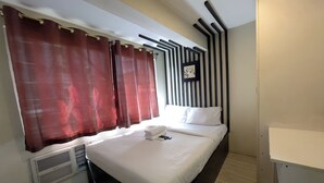 A hotel room with a large bed, a bedside table with a phone, a wall-mounted air conditioner, and a window with red curtains. - Jardin Oriental Suites Ubelt Manila (Manila)