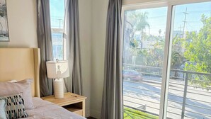 Ein Schlafzimmer mit einem großen Bett, einem Nachttisch mit Lampe und Blick auf eine Straße durch eine Glastür.