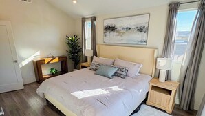 Ein Schlafzimmer mit einem großen Bett, einem Nachttisch, einer Lampe, einem Fenster mit Vorhängen und einem Gemälde an der Wand.