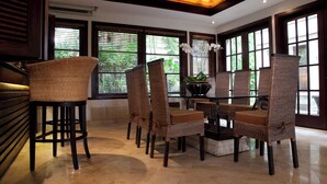 A dining area with wicker chairs and a glass table, large windows with blinds, and a ceiling fan. - Villa Amani (Tabanan)