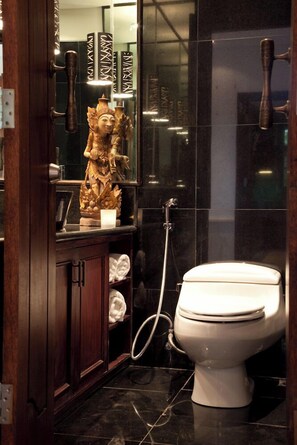 A bathroom with a white toilet, a wooden cabinet, a decorative statue, and a mirror with a black tiled wall. - Villa Amani (Tabanan)