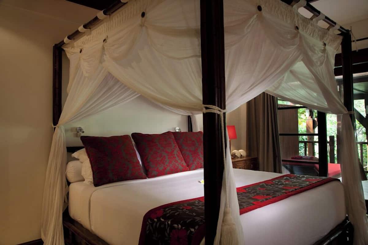 A four-poster canopy bed with red and white bedding, a nightstand with a lamp, and a view of greenery outside.