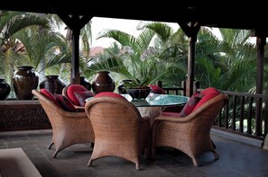 Un espace extérieur aménagé, abrité et doté de chaises en osier et d’une table en verre, entouré de grands vases et de palmiers.
