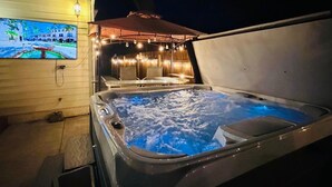 A hot tub with a view of a street scene, a covered seating area with string lights, and a television screen displaying a landscape. -   Heated Pool Hot Tub PicklebalMovieTheaterPoolTable GameR Golf (Des Moines)