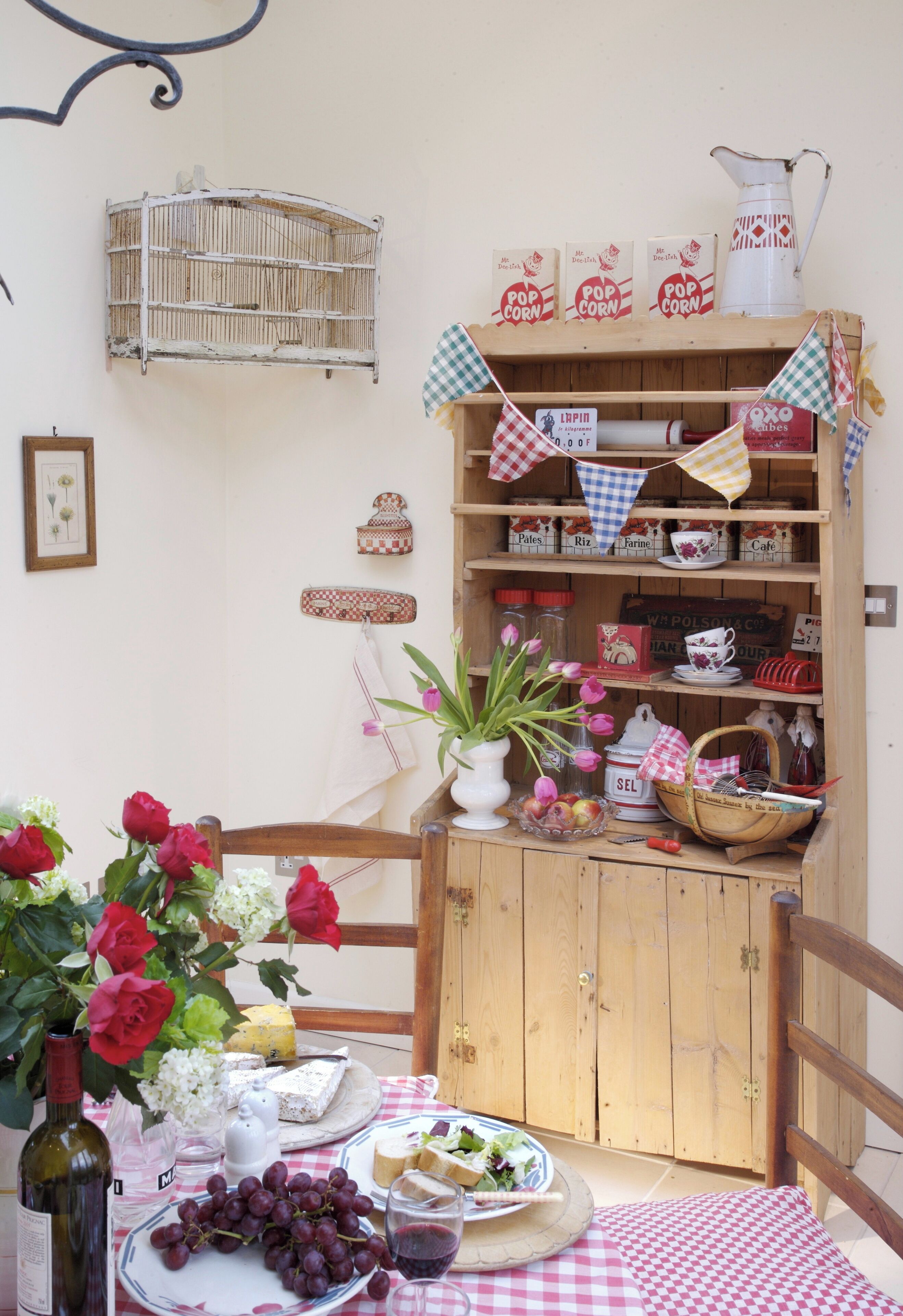 A rustic dining area with a wooden table set for a meal, surrounded by shelves stocked with kitchenware and a birdcage on the wall.