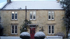 Een stenen gebouw met een rode deur, twee grote sneeuwbemande struiken, en een bank ervoor.