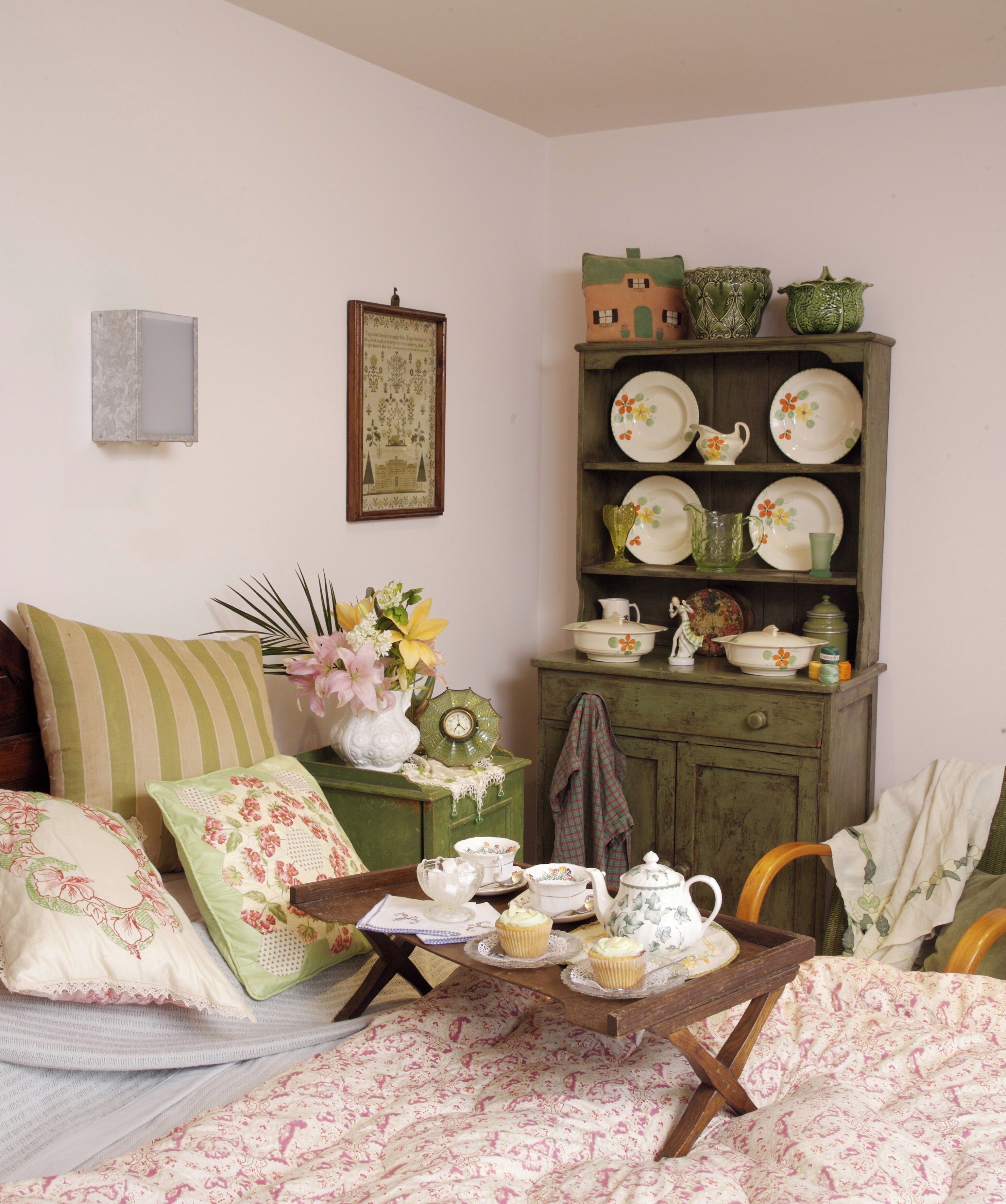 A cozy room with a bed, a wooden cabinet with dishes, a small table with tea set, and floral pillows.