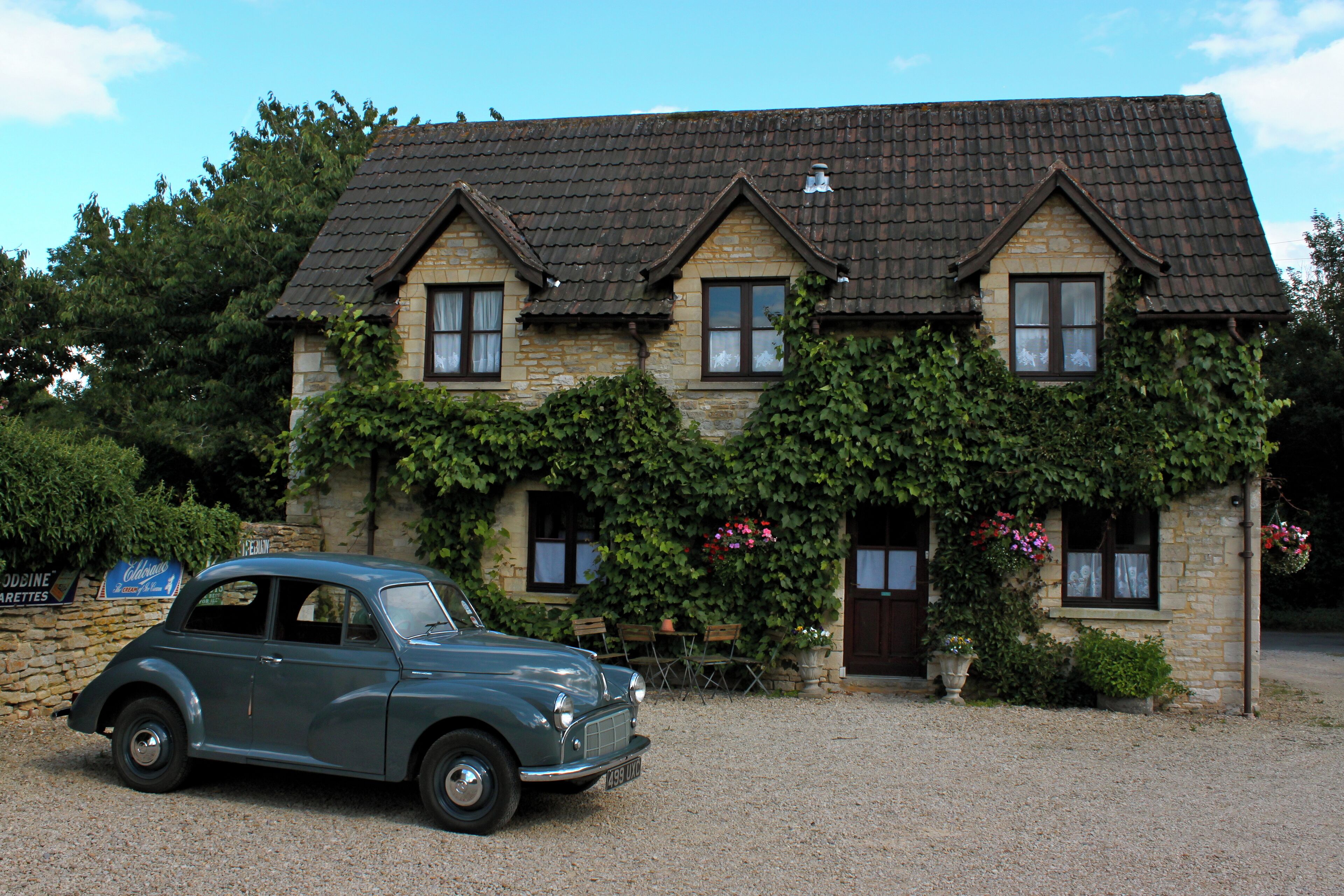 A stone building with a tiled roof, ivy-covered walls, and a vintage car parked in front.