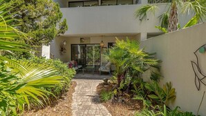 A courtyard with a pathway, surrounded by lush greenery and a seating area with chairs.