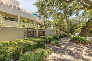 Um edifício moderno com fachada branca, entrada gradeada e um jardim bem cuidado com arbustos e árvores diversos.