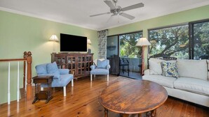 A living room with a wooden floor, a round coffee table, a sofa, two armchairs, a TV, and a ceiling fan.
