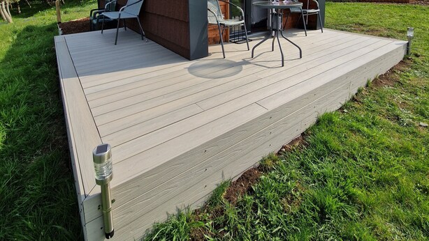 Una cabaña moderna de madera, en parte abierta, con una terraza amueblada con sillas y una mesa, ubicada en un área cubierta de césped con colinas al fondo.