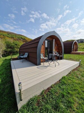 A modern, semi-open wooden cabin with a deck, furnished with chairs and a table, set in a grassy area with hills in the background.