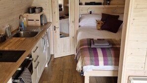 Un dormitorio acogedor y compacto con techo y paredes de madera, una cama con manta escocesa y una pequeña kitchenette con lavamanos y armarios.