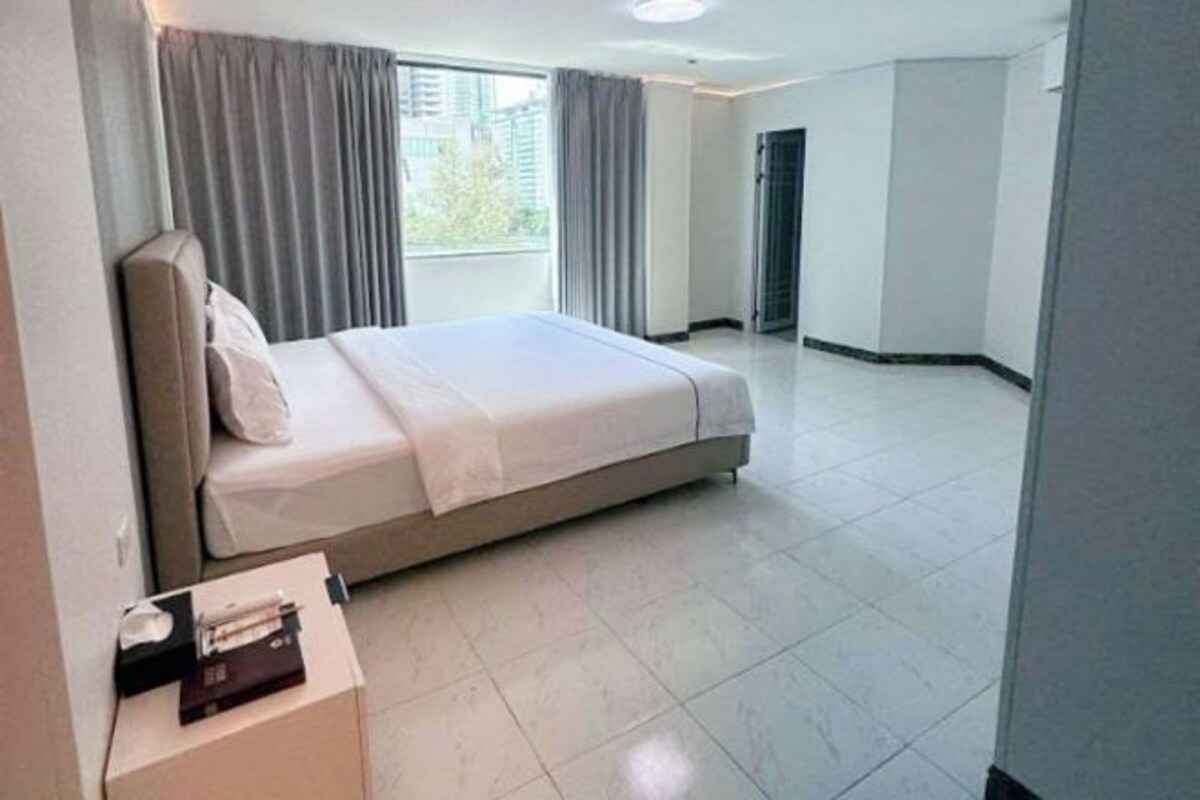 A hotel room with a bed, a nightstand, a window with curtains, and a view of buildings outside.