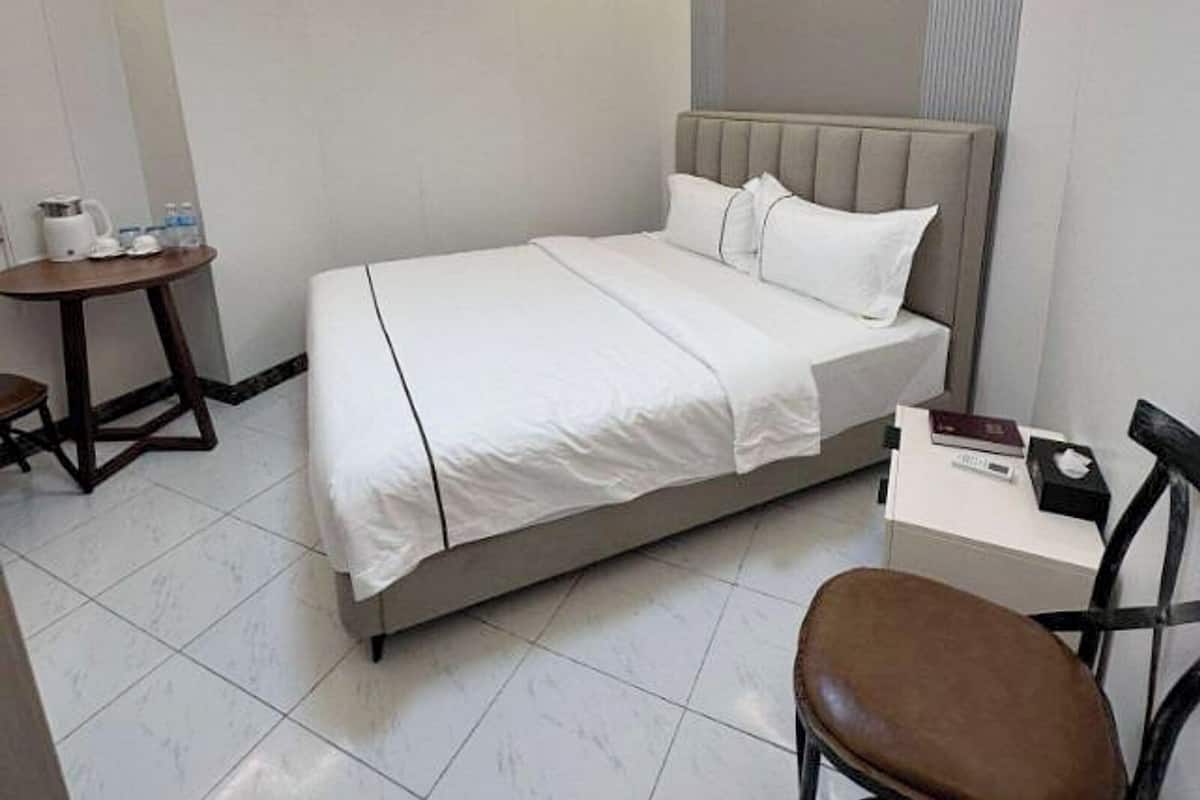 A hotel room with a bed, a small table with a kettle and water bottles, a chair, and a bedside table with a book and tissues.