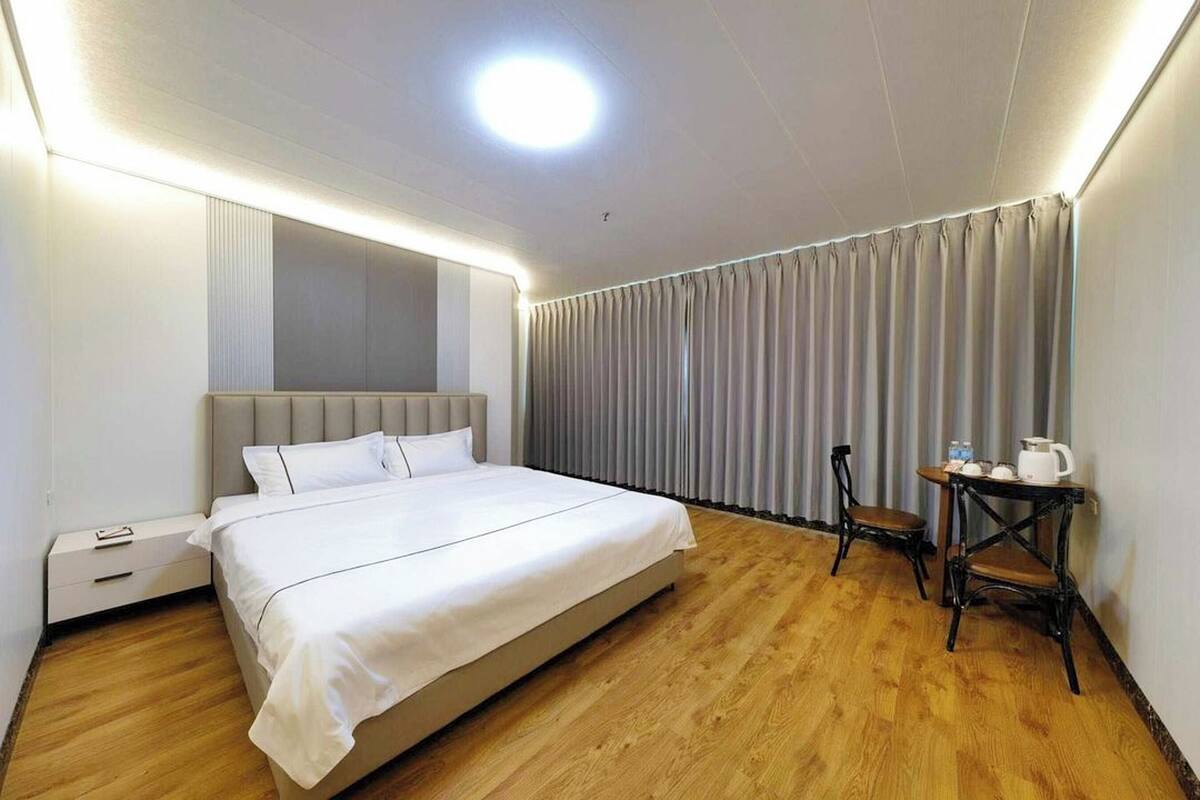 A hotel room with a large bed, a bedside table with a kettle, a chair, and a curtain-covered window.