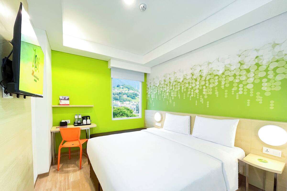 A hotel room with a large bed, a desk with a chair, a window with a view, and a wall with a floral pattern.