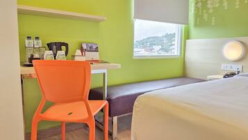 A hotel room with a bed, a desk with a chair, a window with a view, and a bedside table with a lamp.