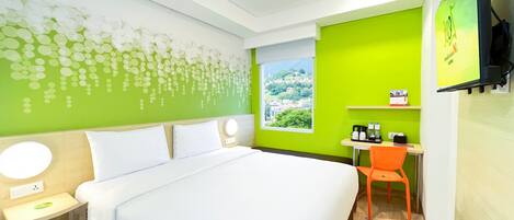 A hotel room with a bed, a desk, a chair, a window with a view, and a wall with a green mural.
