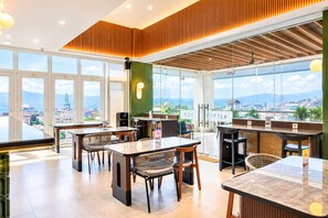 Un restaurante moderno con amplios ventanales que ofrecen vistas de la ciudad, paneles de madera en el techo y diversas opciones de asientos.