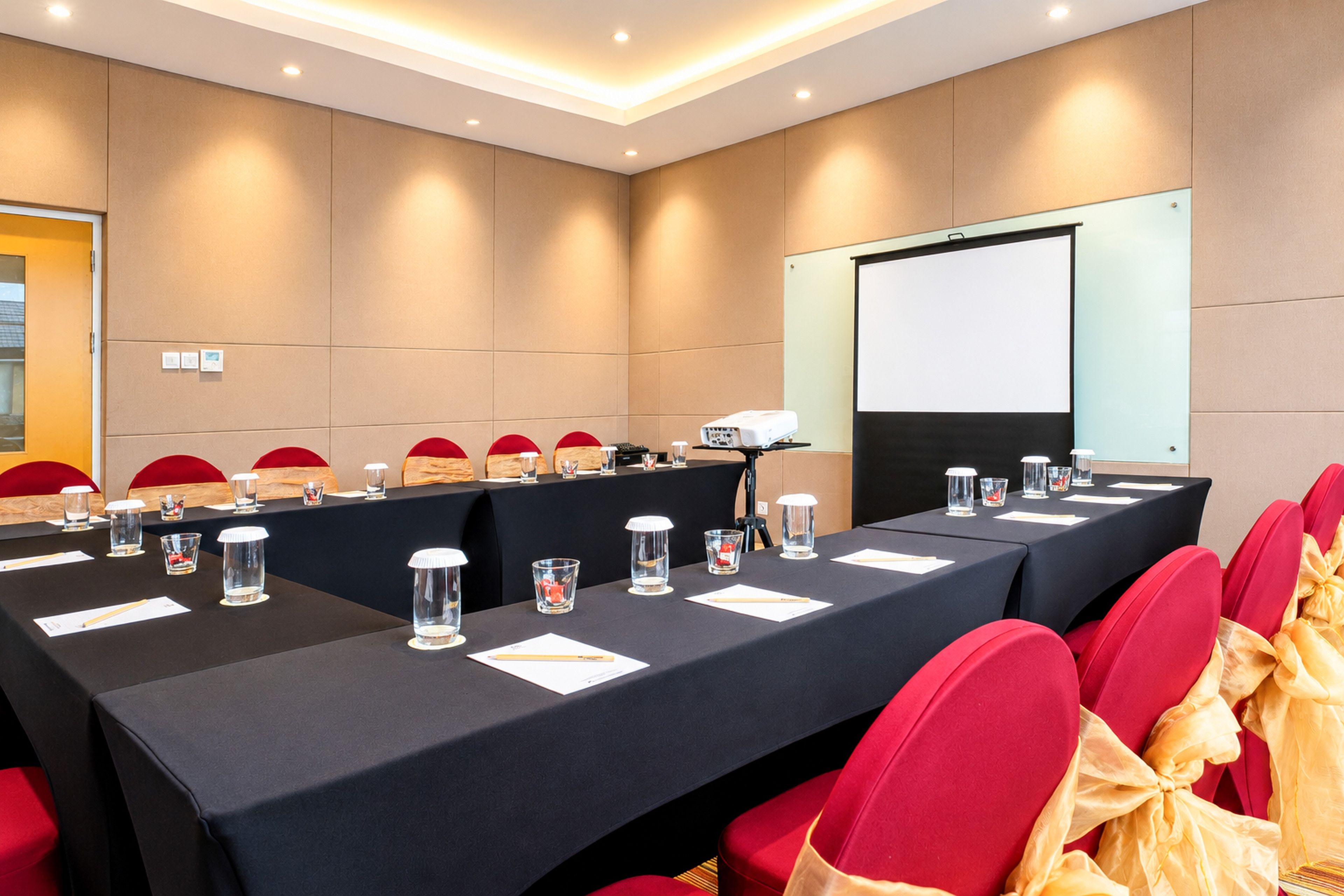 Un salón de conferencias con una mesa larga, sillas rojas y una pantalla de proyector.