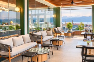 Un bar en la azotea con mesas y sofás al aire libre, con vistas a un paisaje urbano y montañas a lo lejos.