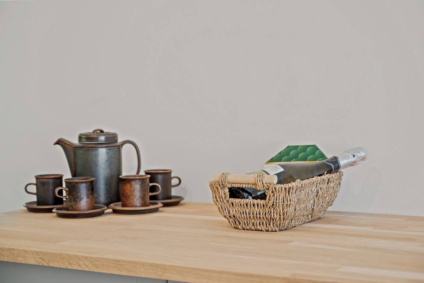A wooden table with a rustic tea set and a woven basket containing a bottle and a green object.