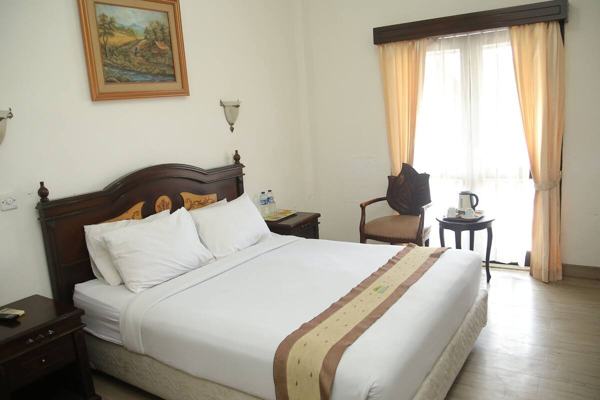 Una cama bien hecha con sábanas blancas, un cabecero de madera y una almohada decorativa. Hay una mesita con una tetera y tazas, y una silla con cojín. Un cuadro cuelga de la pared sobre la cama.